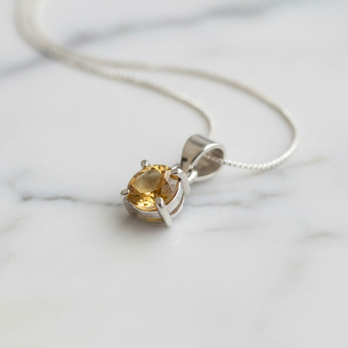 Astral Citrine Round Necklace - Sterling Silver, a silver pendant necklace with an oval-cut yellow citrine gemstone in a prong setting, resting on a white marble surface.
