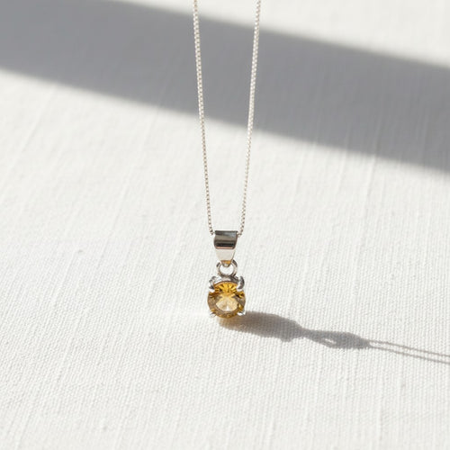 Astral Citrine Round Necklace - Sterling Silver, a delicate silver chain necklace with a small bezel-set round yellow citrine pendant, displayed on a textured white surface with soft shadows.