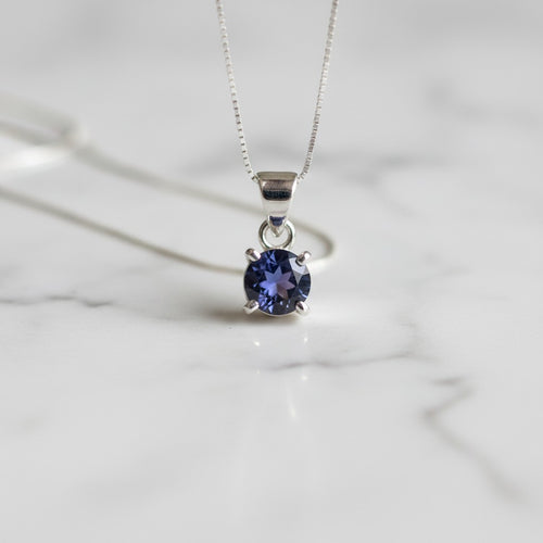 Astral Iolite Round Necklace - Sterling Silver, a silver pendant necklace with a round-cut deep blue iolite gemstone, resting on a white marble surface.