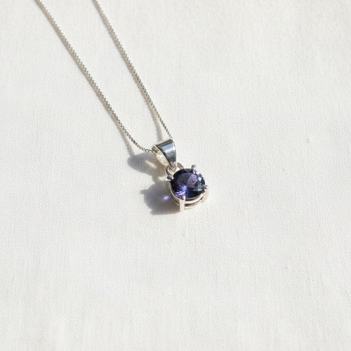 Astral Iolite Round Necklace - Sterling Silver, a silver-toned necklace with a bezel-set oval deep blue iolite pendant, resting on a textured off-white surface.