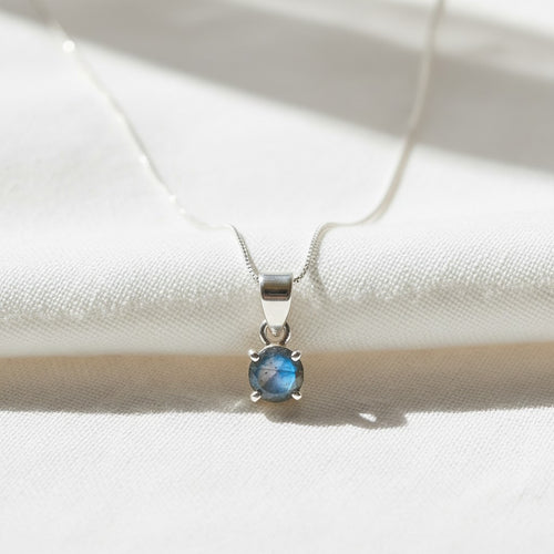Astral Labradorite Round Necklace - Sterling Silver, a silver pendant necklace featuring a round blue gemstone in a four-prong setting, resting on a textured white fabric background with soft shadows.