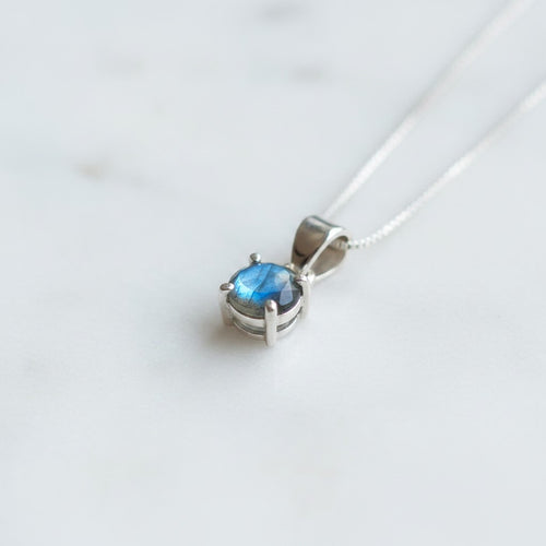 Astral Labradorite Round Necklace - Sterling Silver, a silver pendant necklace featuring a small oval blue gemstone in a prong setting, resting on a light marble surface.