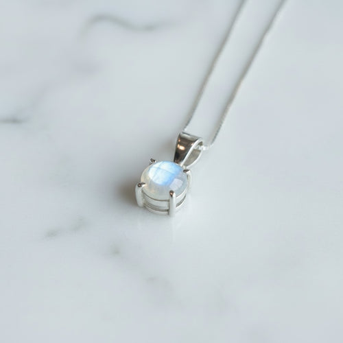 Astral Moonstone Round Necklace - Sterling Silver, a silver pendant necklace featuring an oval moonstone gemstone with a soft blue and white iridescent glow, resting on a white marble surface.