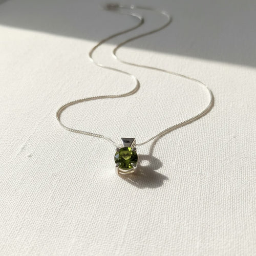 Astral Peridot Round Necklace - Sterling Silver, a silver chain necklace with a small pendant featuring a round green peridot gemstone, displayed on a textured white fabric surface with soft shadows.