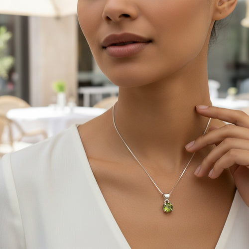 Astral Peridot Round Necklace - Sterling Silver, a close-up lifestyle shot of a woman wearing a delicate silver chain necklace with a small round green peridot pendant, set against a softly blurred cafe background.