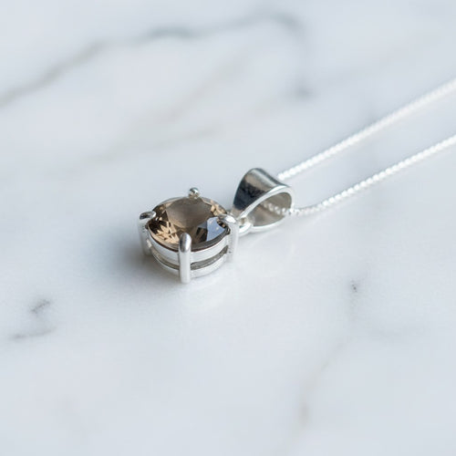Astral Smoky Quartz Round Necklace - Sterling Silver, a silver pendant necklace with a round-cut smoky brown quartz gemstone, resting on a white marble surface.