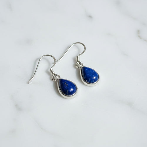 Athena Lapis Lazuli Teardrop Earrings - Sterling Silver, a pair of silver dangle earrings with teardrop-shaped deep blue lapis lazuli stones, displayed on a white marble surface.