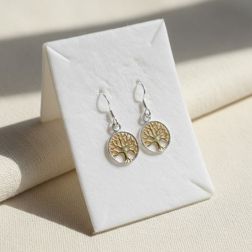 Auryn Tree of Life Drop Earrings - Sterling Silver & Gold Plate, a pair of dangling tree-of-life earrings displayed on a white jewelry stand, with a silver-tone fishhook back and circular dangle with a gold-tone tree design set against a cream textured background.