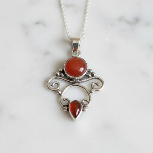 Awen Carnelian Necklace - Sterling Silver, a silver pendant necklace featuring two red cabochon and teardrop gemstones, displayed on a white marble surface.