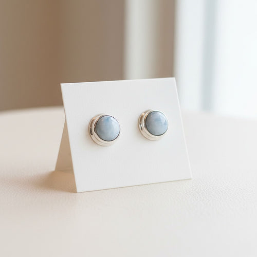 Marina Larimar Round Stud Earrings - Sterling Silver, a pair of round stud earrings with pale blue-grey marbled stones in silver bezel settings, displayed on a small white stand.