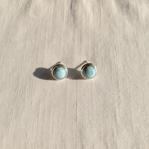 Marina Larimar Round Stud Earrings - Sterling Silver, a pair of round stud earrings with light blue-green marbled stones in silver bezel settings, resting on a textured beige linen surface.