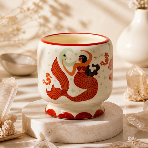 Paddywax A Dopo Candle - Mermaid & Moon, Ginger & Teak, a ceramic candle holder featuring a whimsical mermaid illustration in red, orange, black, and cream, displayed on a round white marble riser with crystals and dried botanicals in the background.