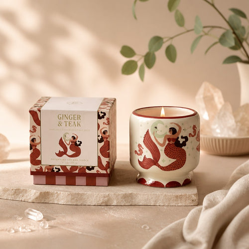 Paddywax A Dopo Candle - Mermaid & Moon, Ginger & Teak, a Ginger & Teak scented candle in a cream-coloured ceramic jar with a red mermaid illustration, lit and shown next to its matching patterned packaging box on a stone slab with warm, soft-focus background props.