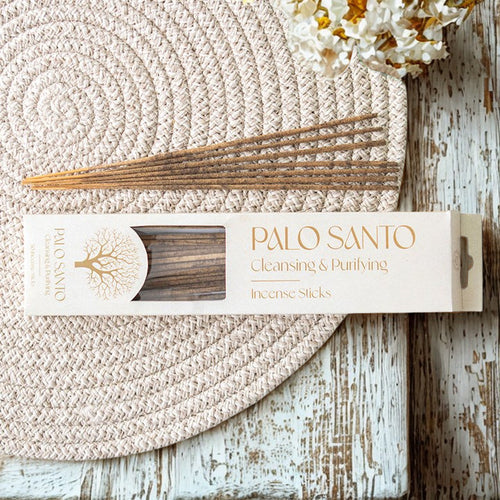 Palo Santo Incense Sticks - Pack Of 30, loose sticks and packaging on a woven placemat with dried white flowers, on a rustic wooden surface.