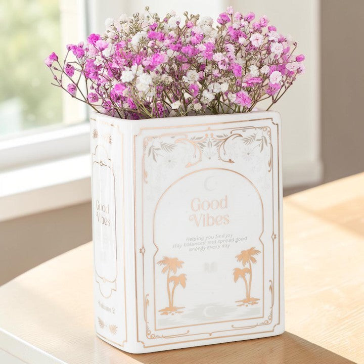 White Book Shaped Vase - Good Vibes, book-shaped ceramic vase filled with pink and white flowers on a wooden table.