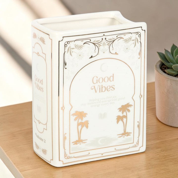 White Book Shaped Vase - Good Vibes, angled view of the white ceramic book-shaped vase showing the spine detail with a small potted succulent plant beside it on a wooden surface.